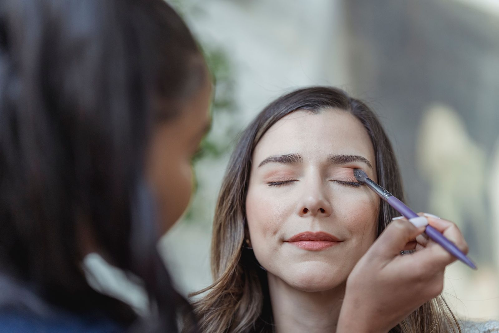 Crop young ethnic female makeup artist using brush while applying eyeshadow on eyelid of client with closed eyes in light beauty salon